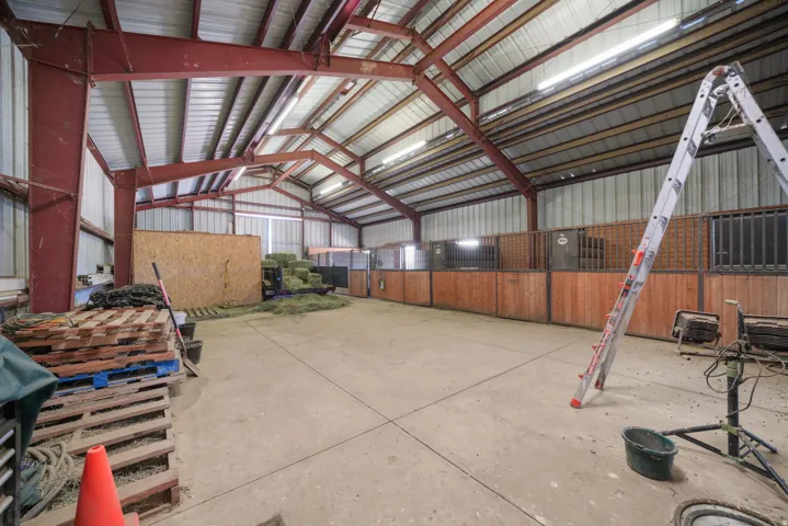 Spacious barn interior featuring robust metal beam construction, high ceilings, concrete flooring, multiple stalls with wood-paneled walls, and fluorescent strip lighting
