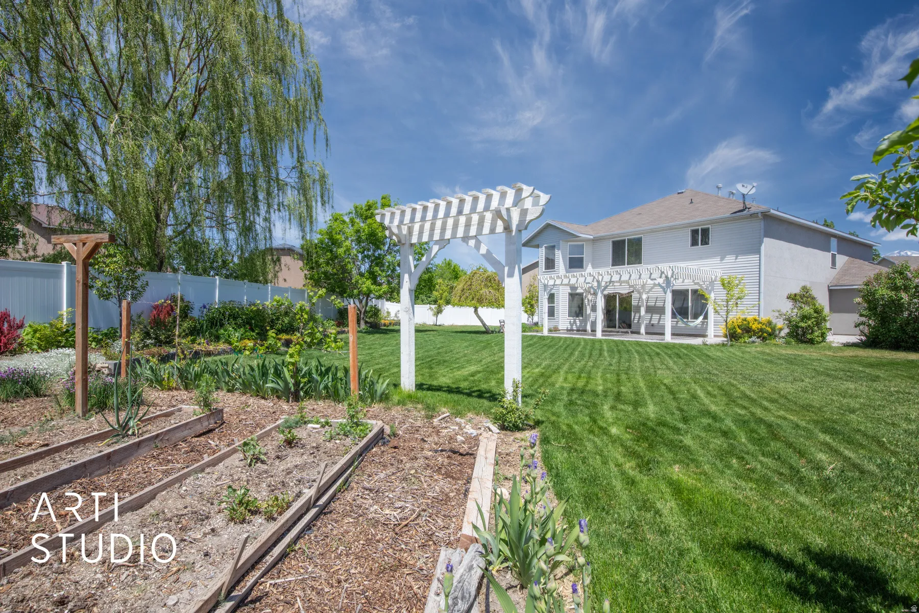 Expansive backyard featuring a manicured lawn, a white pergola-covered patio, raised garden beds, and a white privacy fence