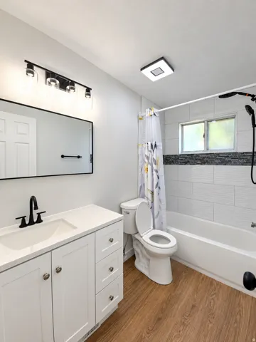 White vanity with a solid surface countertop, matte black faucet, wood-finish flooring, tiled shower surround with accent band, and a window