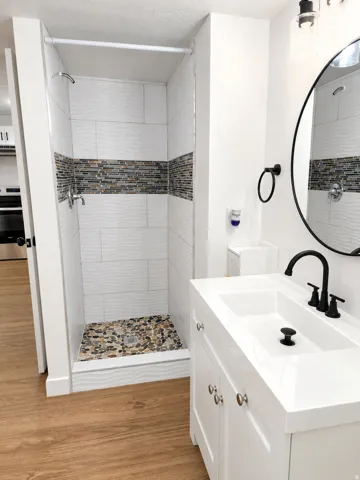 Bathroom featuring a standing shower with textured white tile and a mosaic accent band, pebble-tiled shower floor, white vanity with integrated sink, black faucet, and a round black-framed mirror