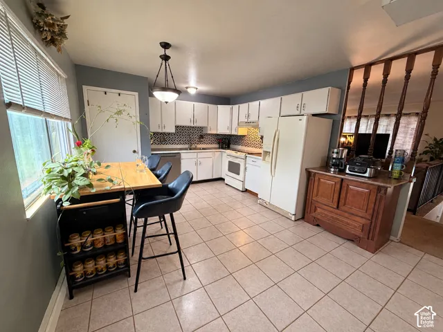Kitchen featuring tile flooring, white cabinetry, a patterned backsplash, white appliances, and a central pendant light fixture