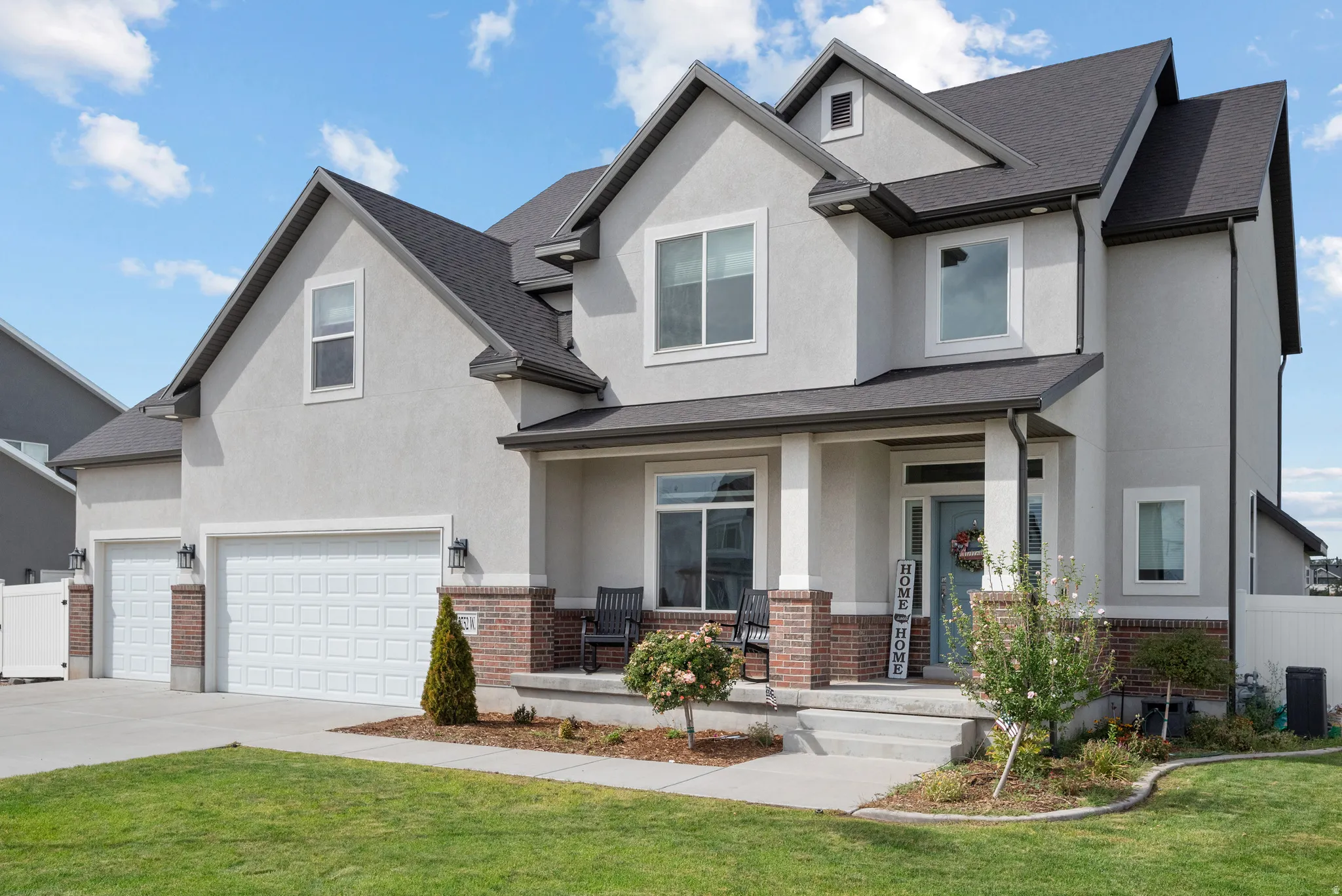 Light-toned stucco exterior with brick accents, front porch with column details, dark shingle roof, and a two-car garage