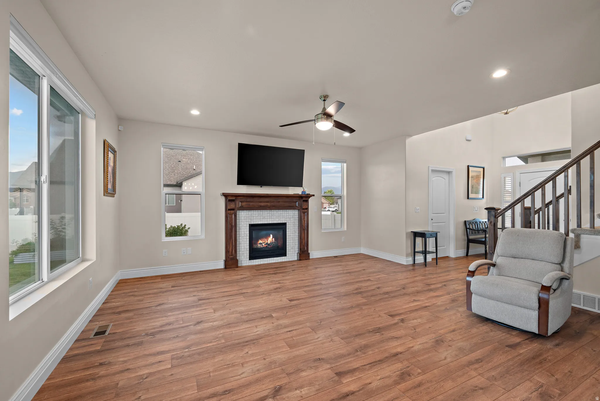Spacious living area featuring wood-finish flooring, a decorative fireplace with tile surround, a ceiling fan, and multiple windows