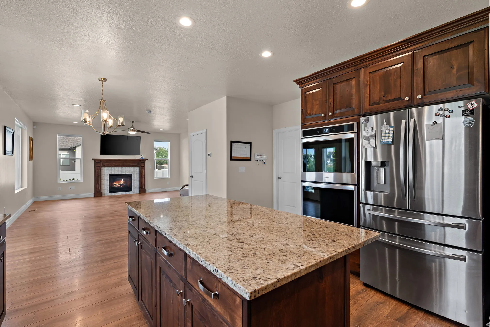 Kitchen island with stone countertop and rich wood cabinetry, stainless steel refrigerator and built-in double ovens, wood-finish flooring, and a living area with a fireplace