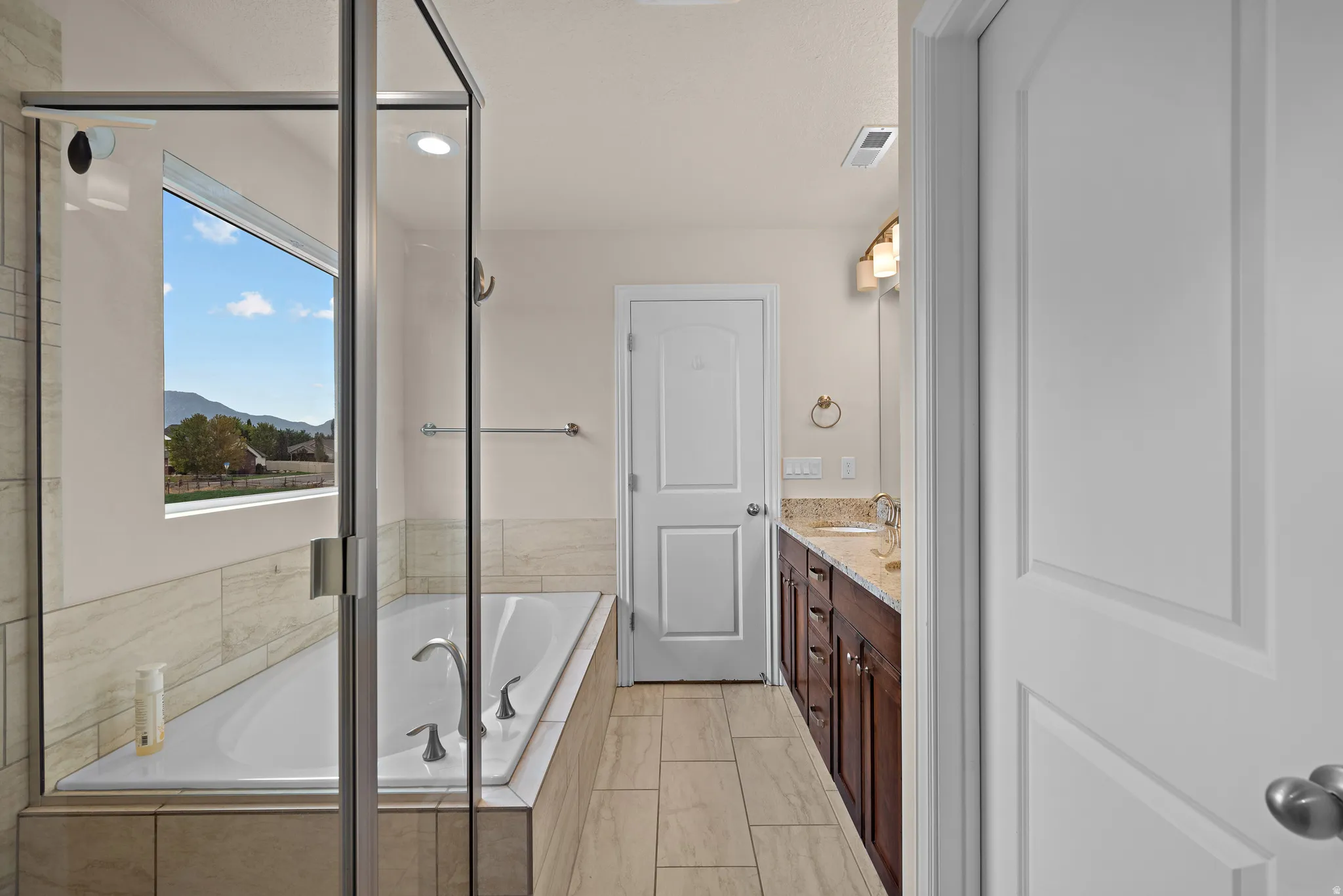Enclosed shower with a built-in tub, a large window, tiled flooring, a wood-finish double vanity, and a light-colored countertop