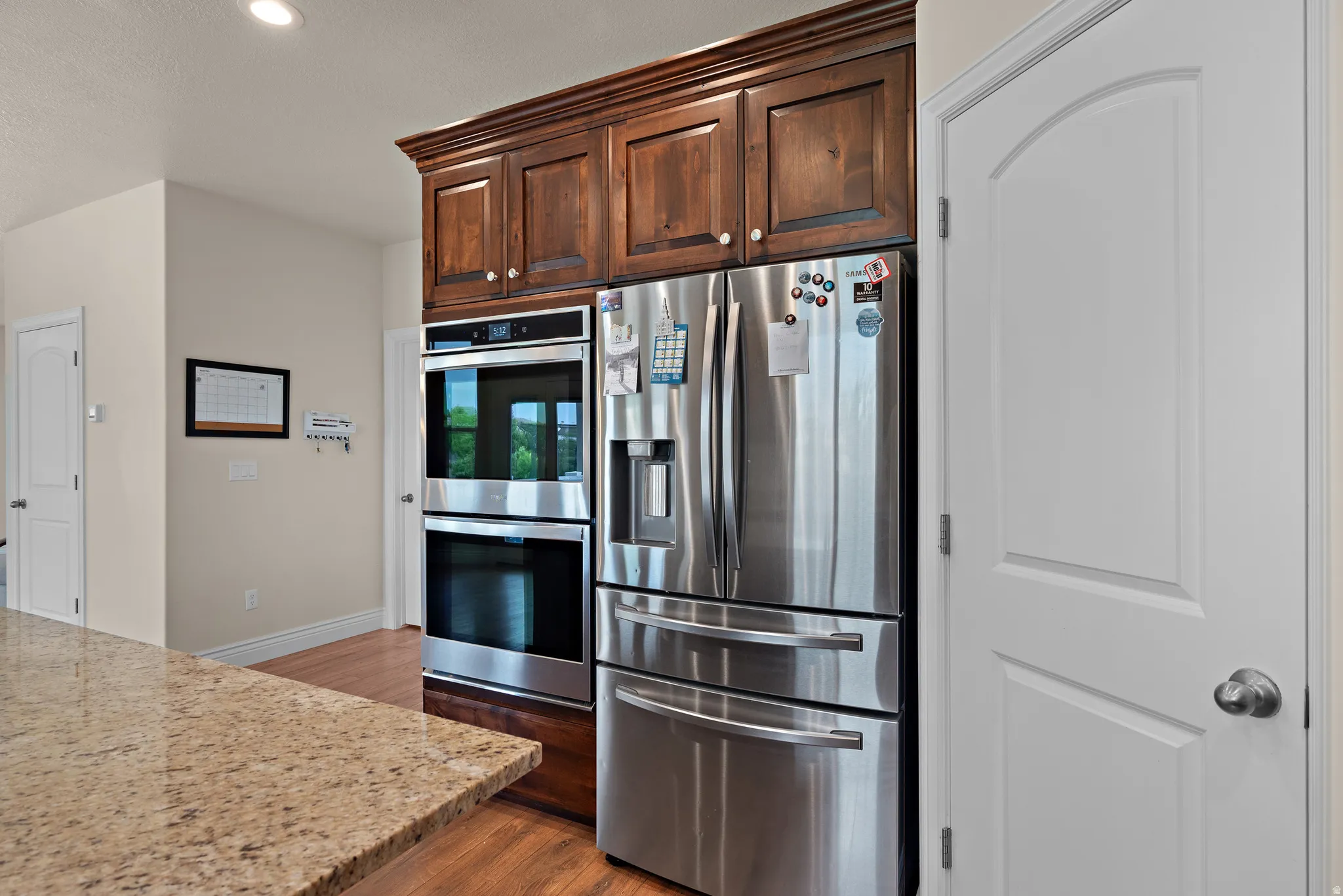 Built-in stainless steel double ovens, stainless steel refrigerator, rich wood cabinetry, light-toned granite countertop, and wood-finish flooring