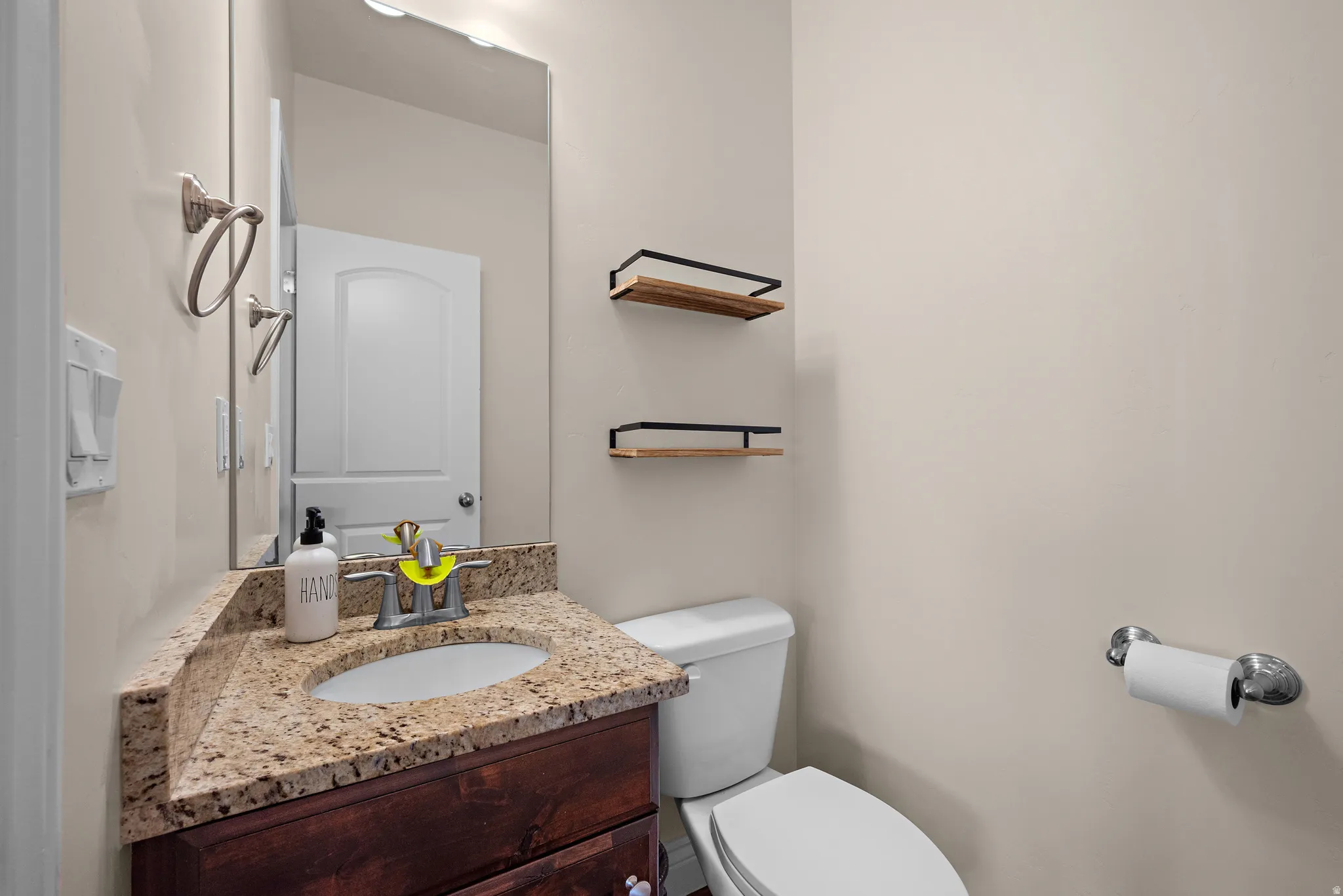 Wood-finish vanity with an undermount sink, granite countertop, wall-mounted mirror, and overhead vanity lighting