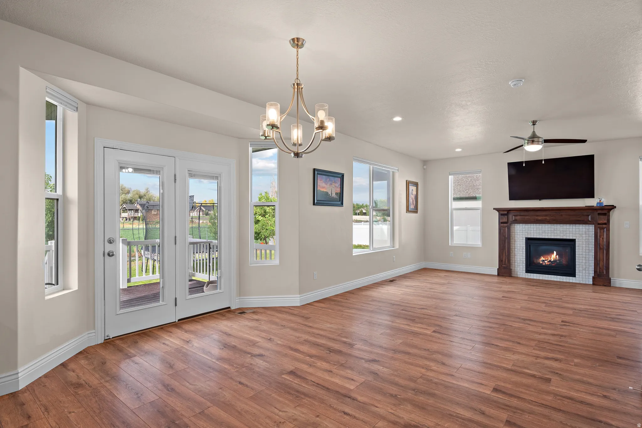 Wood-finish flooring throughout, French doors with direct outdoor access, a tiled fireplace with a substantial wood mantel, multiple windows for natural illumination, and a ceiling fan