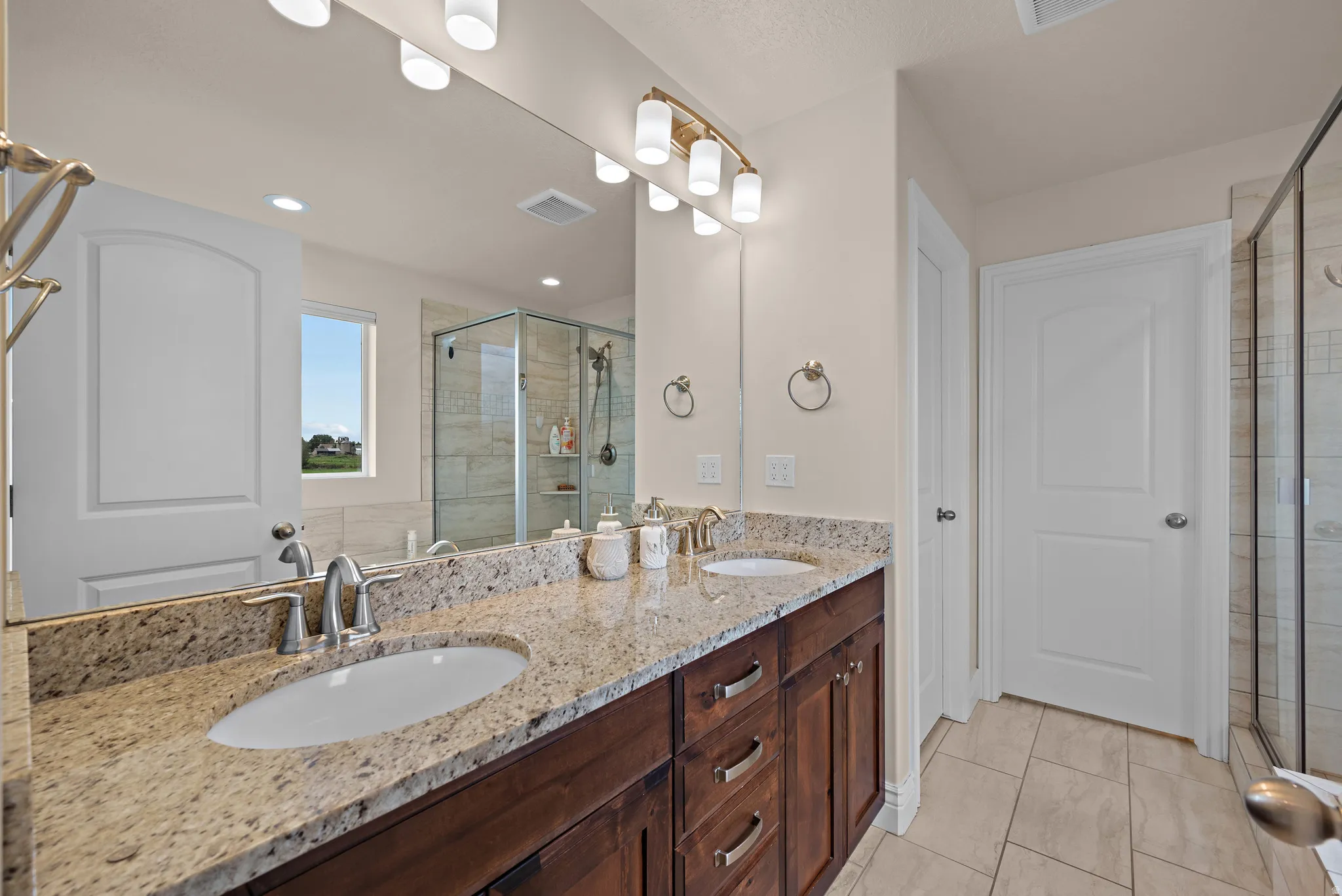 Dual undermount sinks with brushed nickel fixtures, granite countertops, wood-finish cabinetry, a glass-enclosed shower, and tile flooring