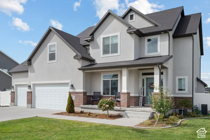 Light-toned stucco exterior with brick accents, front porch with column details, dark shingle roof, and a two-car garage