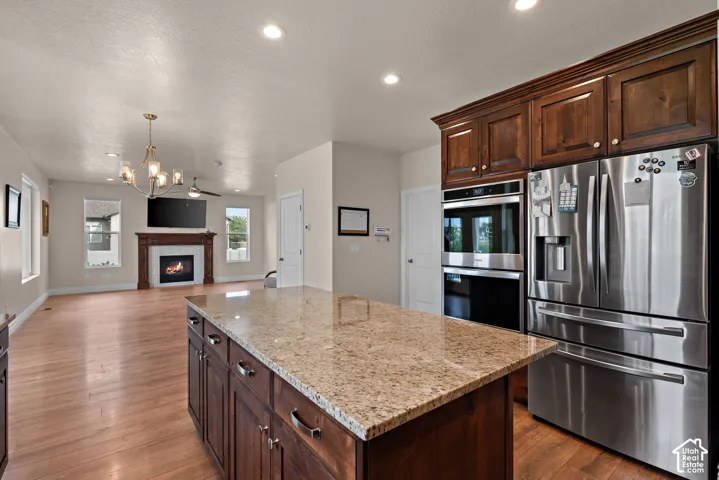 Kitchen island with stone countertop and rich wood cabinetry, stainless steel refrigerator and built-in double ovens, wood-finish flooring, and a living area with a fireplace