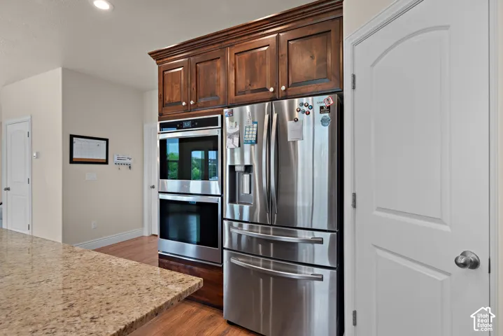 Built-in stainless steel double ovens, stainless steel refrigerator, rich wood cabinetry, light-toned granite countertop, and wood-finish flooring