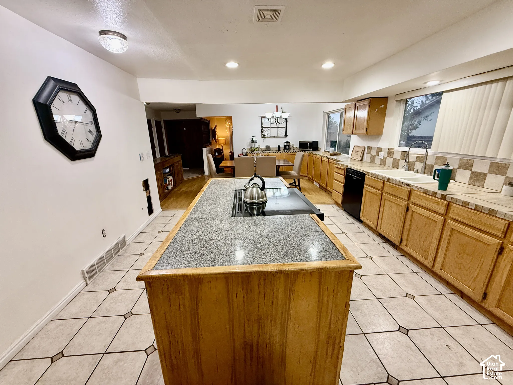 Kitchen with a central island featuring an integrated cooktop, light wood-finish cabinetry, tile countertops, and a checkered tile backsplash