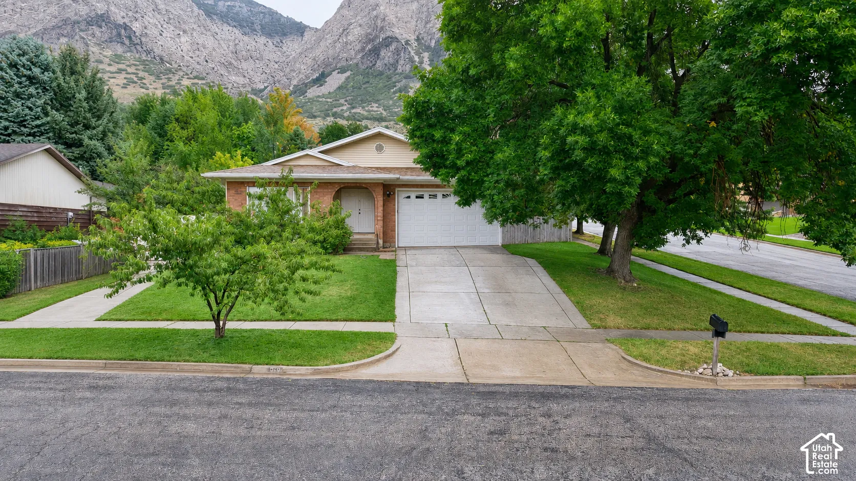 Brick exterior, attached garage, concrete driveway, front lawn with mature trees, and mountainous backdrop