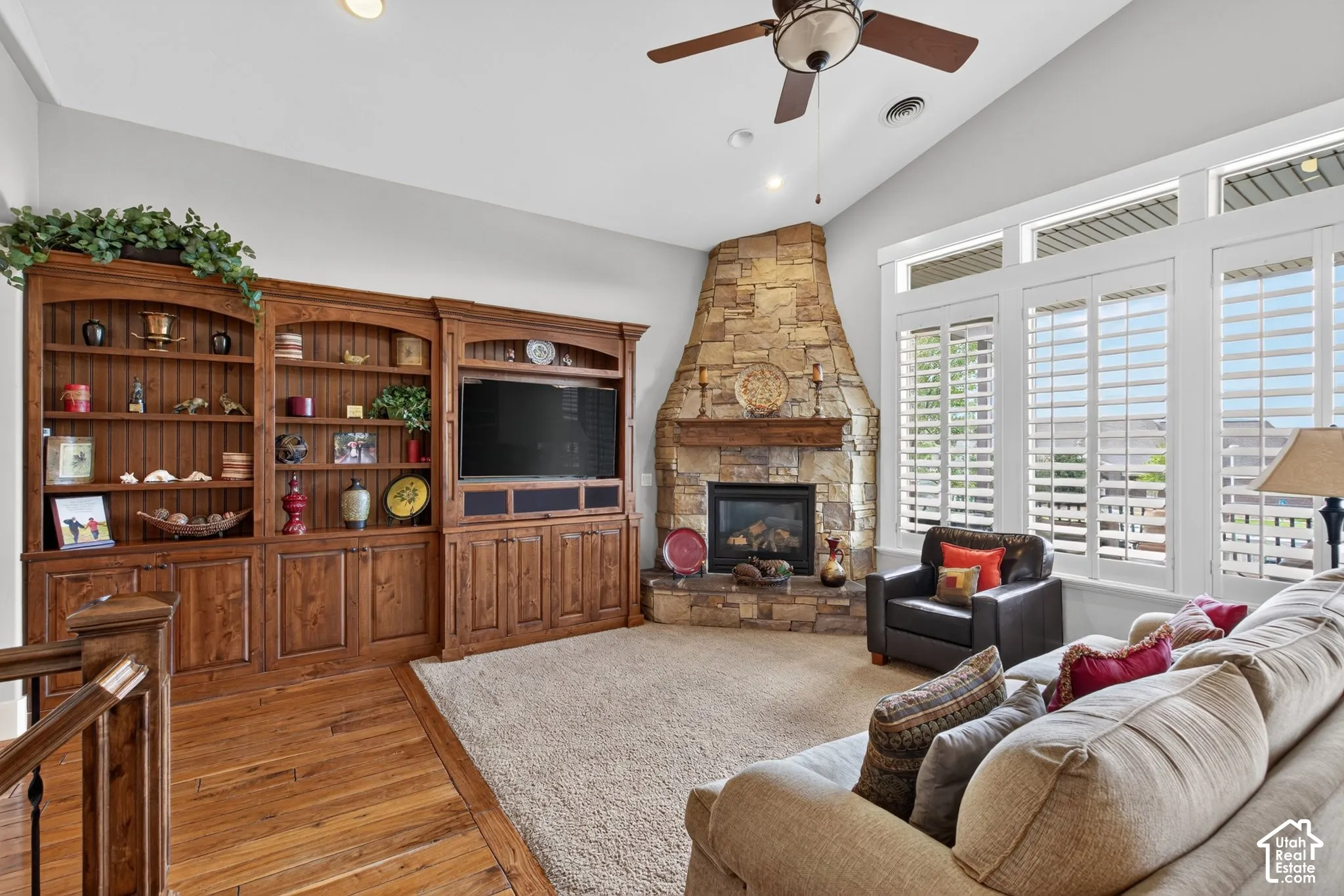 Vaulted ceiling with recessed lighting and ceiling fan, stone fireplace with a wood mantle, built-in custom cabinetry, hardwood flooring, and plantation shutters