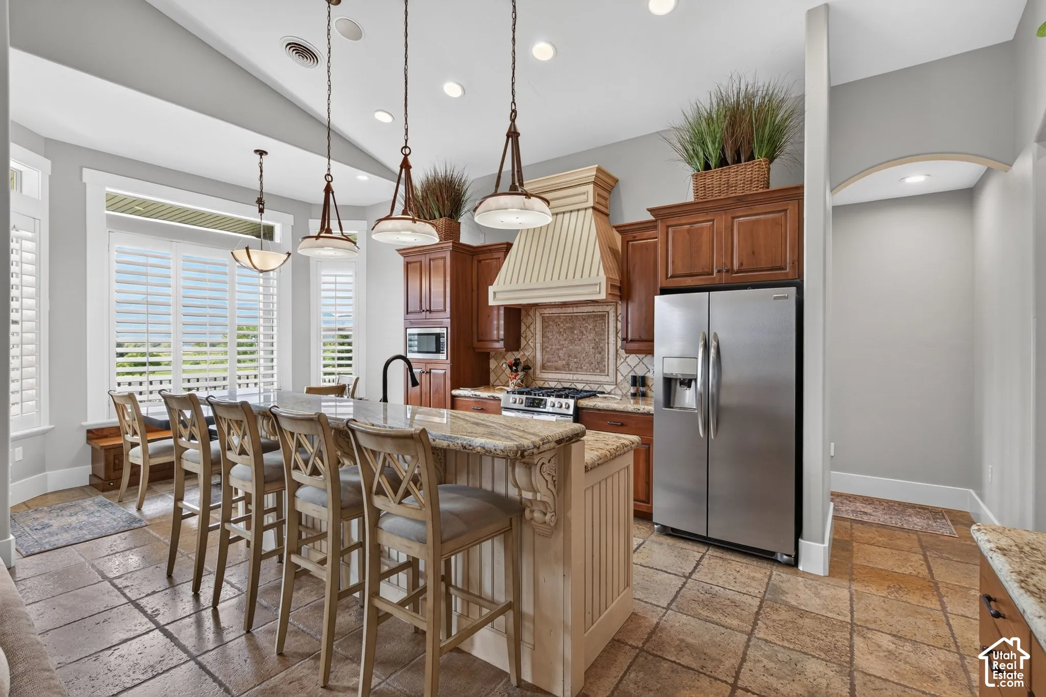 Kitchen featuring a large island with stone countertop, stainless steel appliances, custom wood cabinetry, tiled flooring, and a dining area with window shutters
