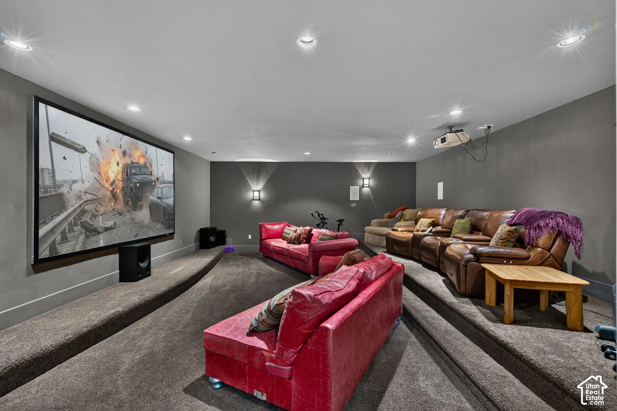 Expansive media room featuring a large projection screen, tiered carpeted flooring, recessed lighting, wall-mounted light fixtures, and a ceiling-mounted projector