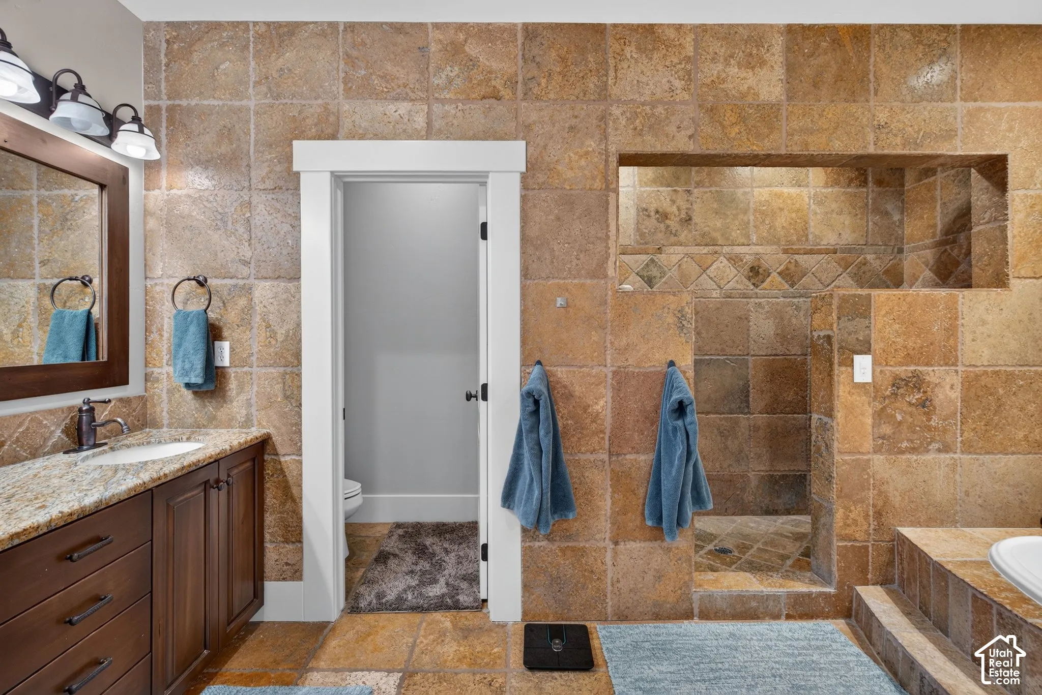 Tiled flooring and walls, a built-in shower niche, a vanity with a granite countertop, and a vessel sink