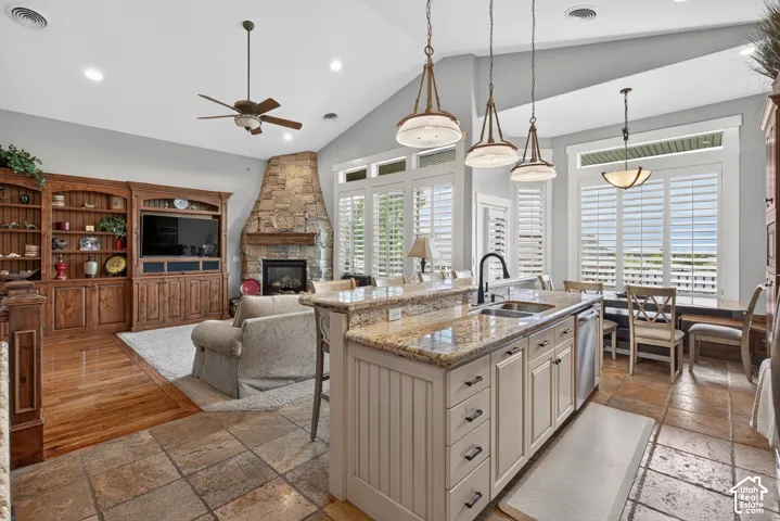 Open-concept living space featuring vaulted ceilings, a stone fireplace, built-in wooden cabinetry, a kitchen island with stone countertops, and plantation shutters