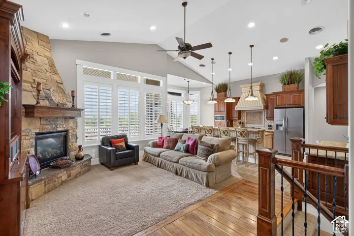 Vaulted ceiling with ceiling fan, stone fireplace with hearth, multiple windows with plantation shutters, wood-finish flooring, and a kitchen featuring pendant lighting and stainless steel appliances