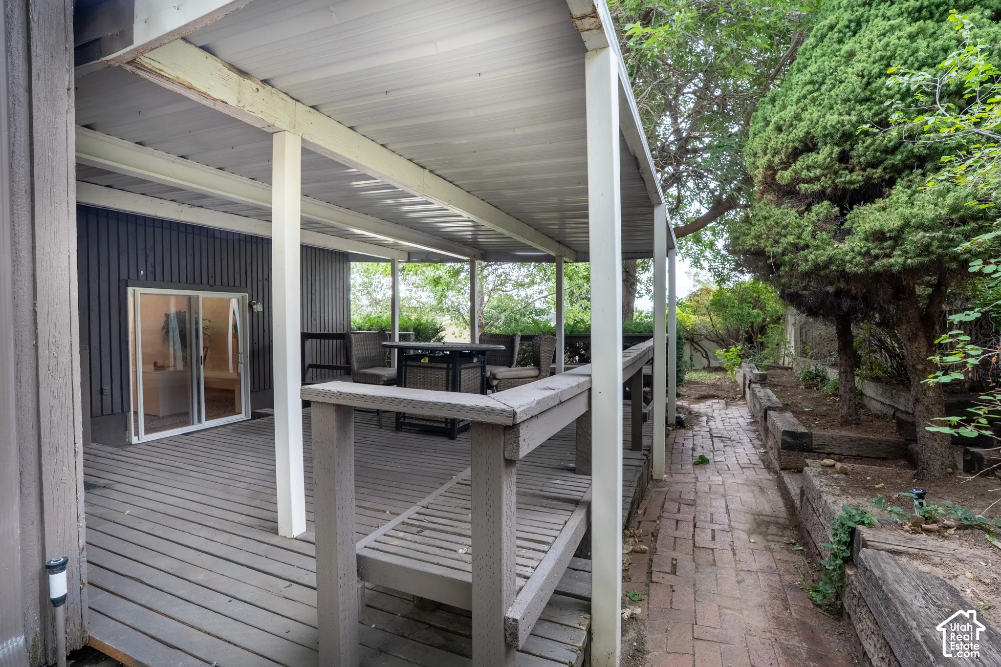 Covered outdoor deck with wood-finish decking and integrated railing, featuring a sliding glass door and a brick paver pathway