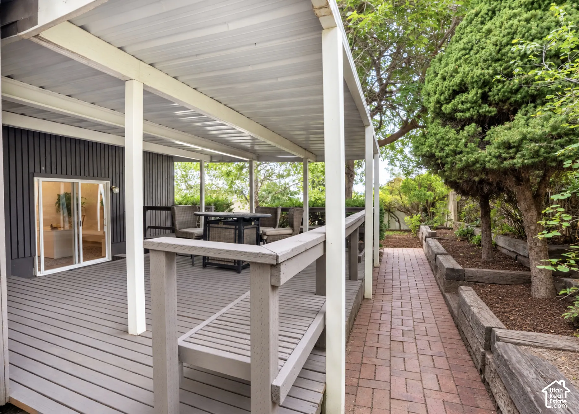 Covered outdoor patio with sliding glass door access to the interior, brick paver walkway, and tiered landscaping with mature trees