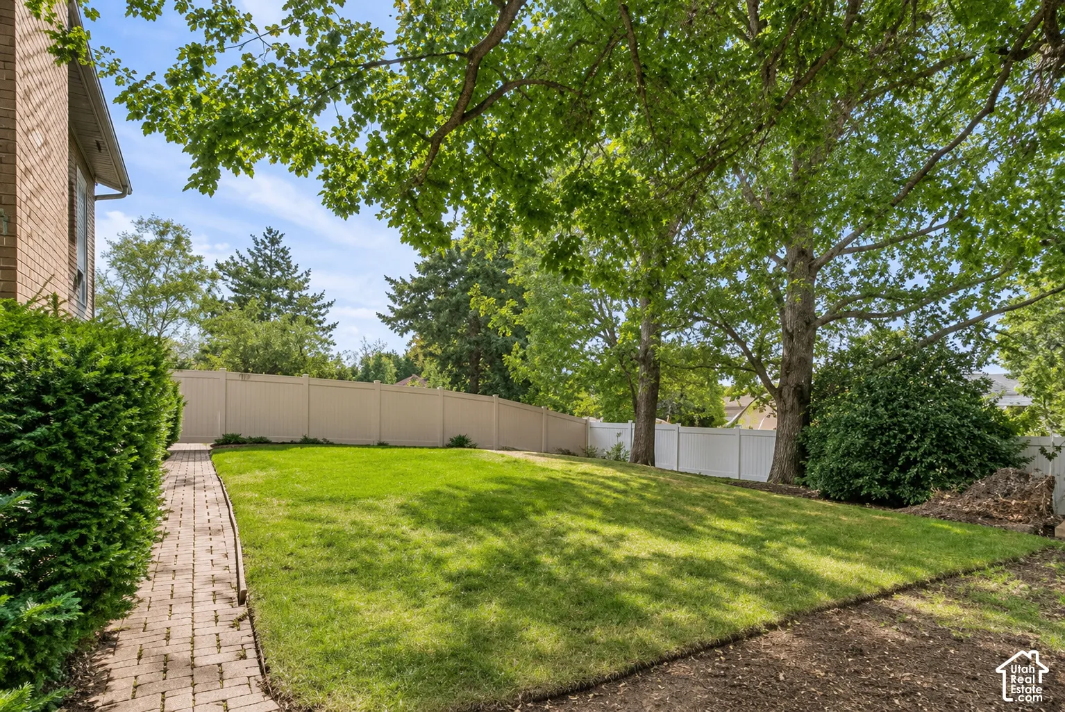 Expansive backyard featuring a lush lawn, mature trees providing shade, a brick paver pathway, and a perimeter fence