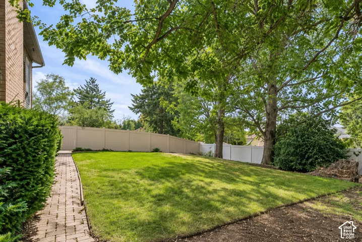 Expansive backyard featuring a lush lawn, mature trees providing shade, a brick paver pathway, and a perimeter fence