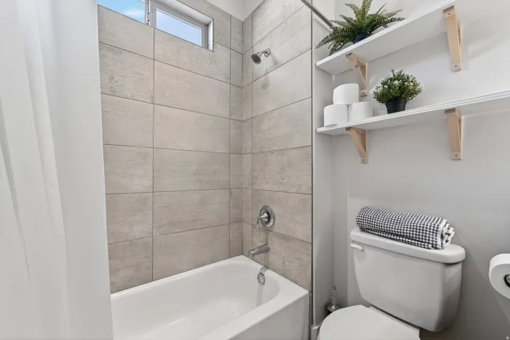 Bathtub with light-toned tiled surround, shower fixture, and an overhead transom window
