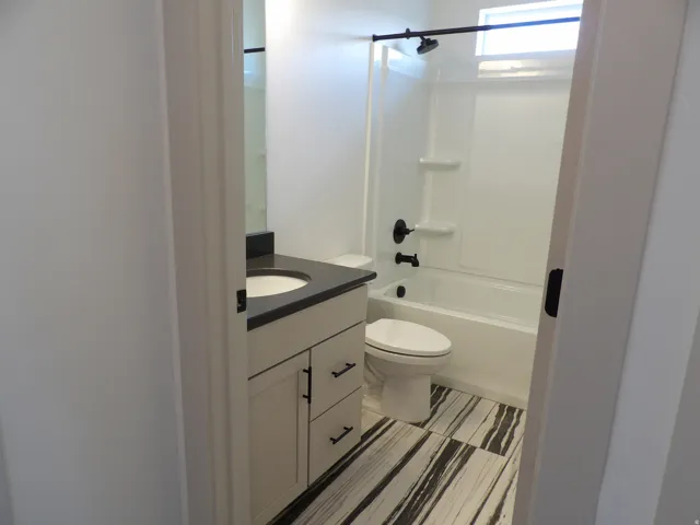 Vanity with a dark countertop and undermount sink, white cabinetry with dark hardware, a bathtub with a shower and dark fixtures, and black and white striped floor tiling