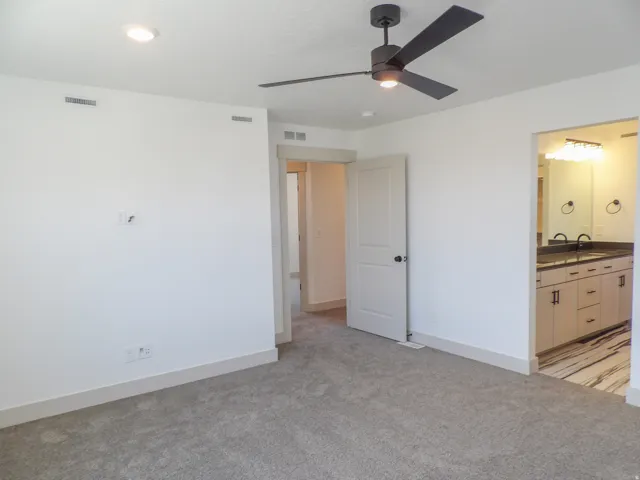 Recessed ceiling lighting, modern ceiling fan, gray carpeting, white walls, and a contrasting bathroom with a dark countertop vanity
