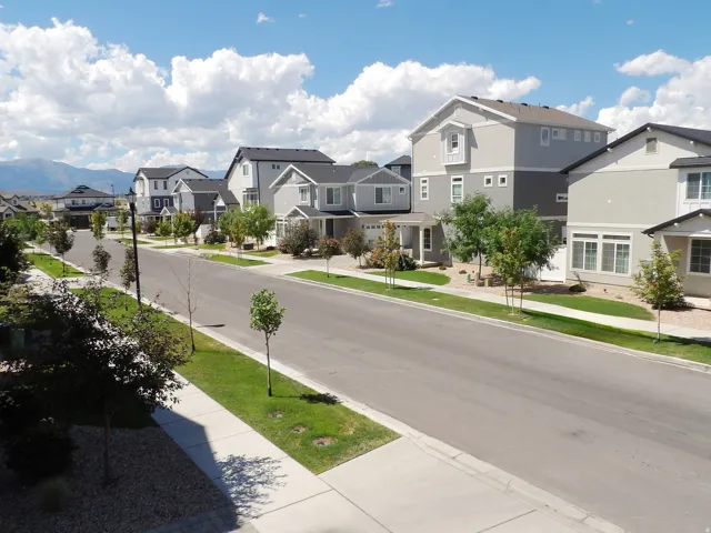 Residential street with sidewalks and green verges, multiple multi-story homes featuring neutral-toned exteriors, and a distant mountain range