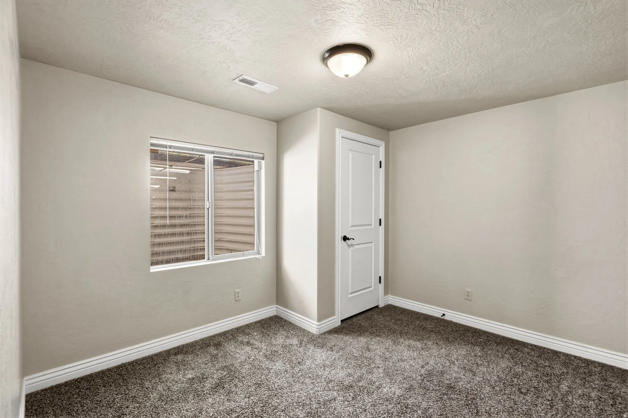 Neutral wall paint, textured ceiling, window with blinds, paneled door with dark hardware, and carpet flooring