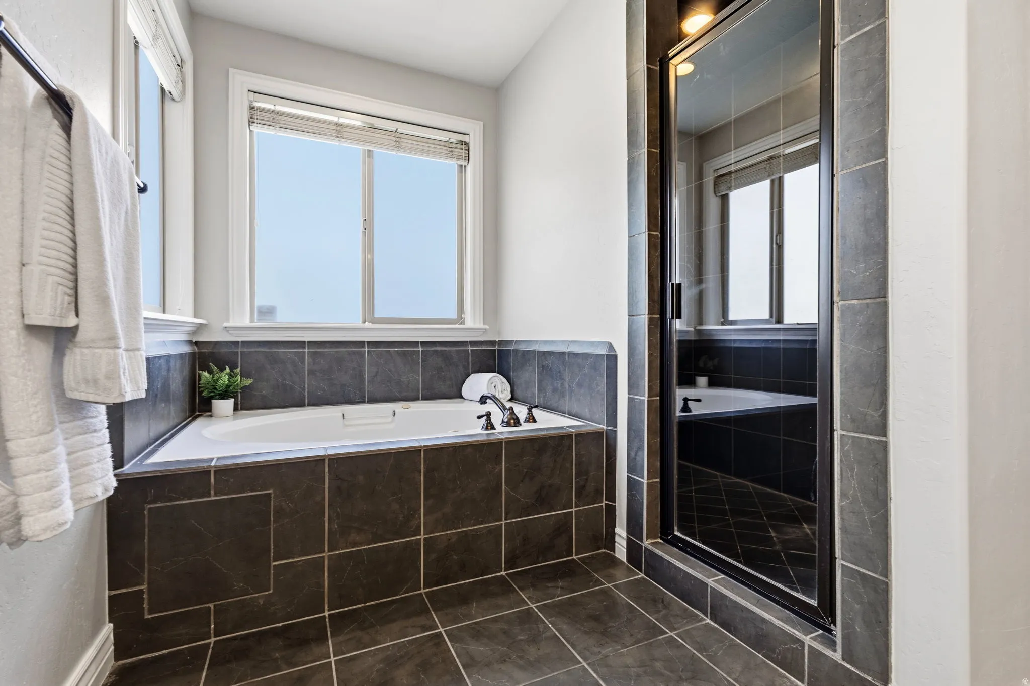 Built-in tub with dark tile surround, dark tile flooring, and a glass-enclosed shower with dark framing