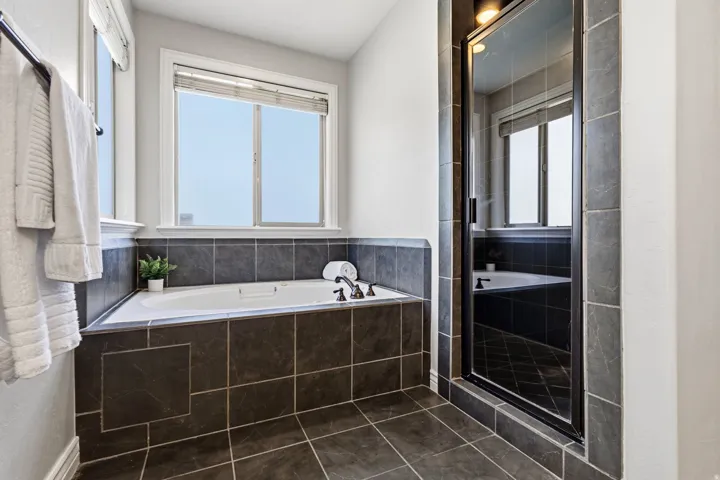 Built-in tub with dark tile surround, dark tile flooring, and a glass-enclosed shower with dark framing