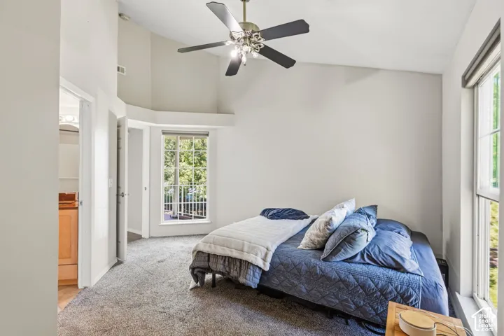 Vaulted ceiling with a ceiling fan, carpeted flooring, large window, and direct access to a bathroom featuring a vanity with a light wood-finish cabinet