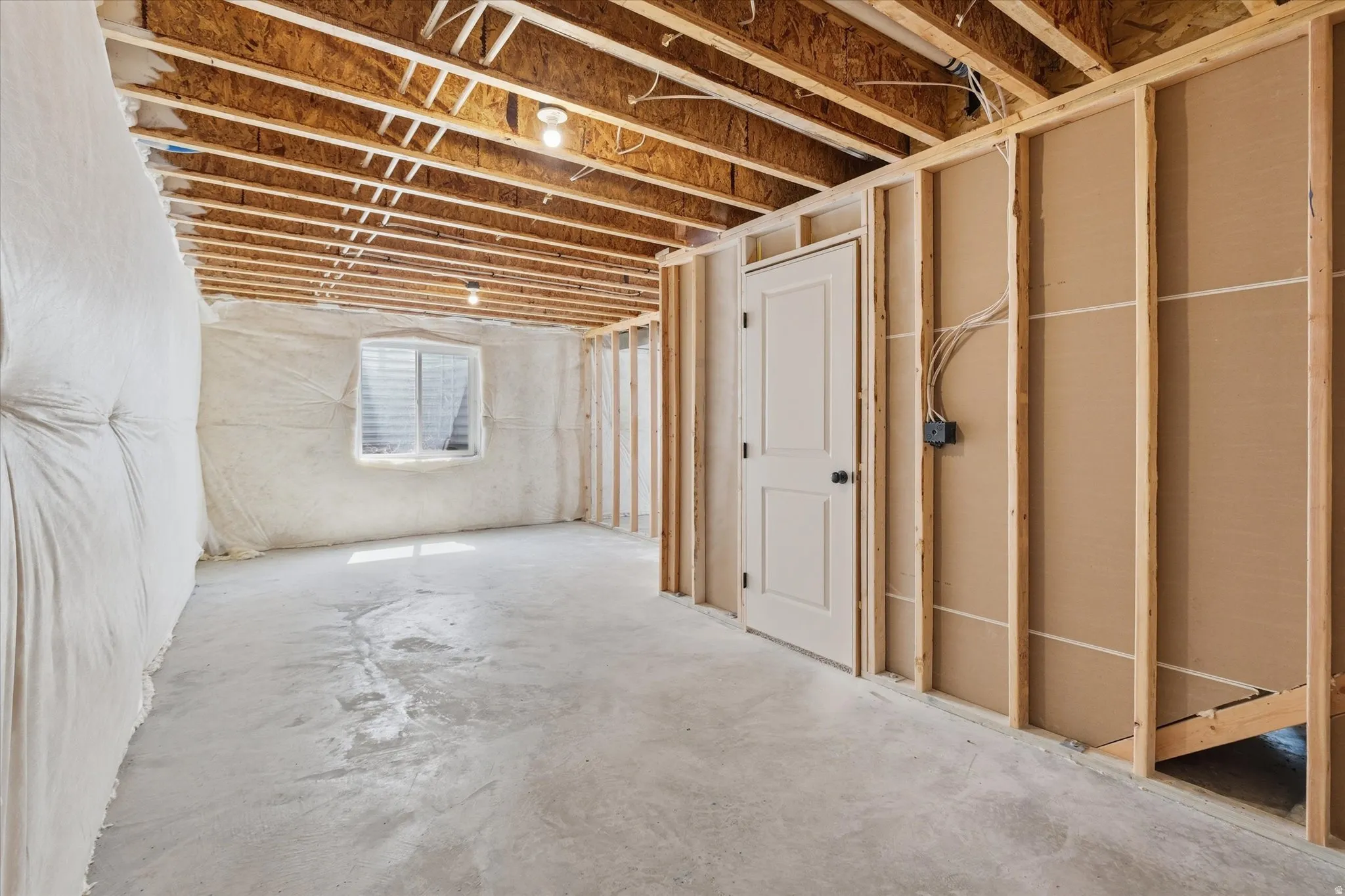 Unfinished basement area featuring exposed ceiling joists, concrete flooring, perimeter insulation, a white interior door, and a single window