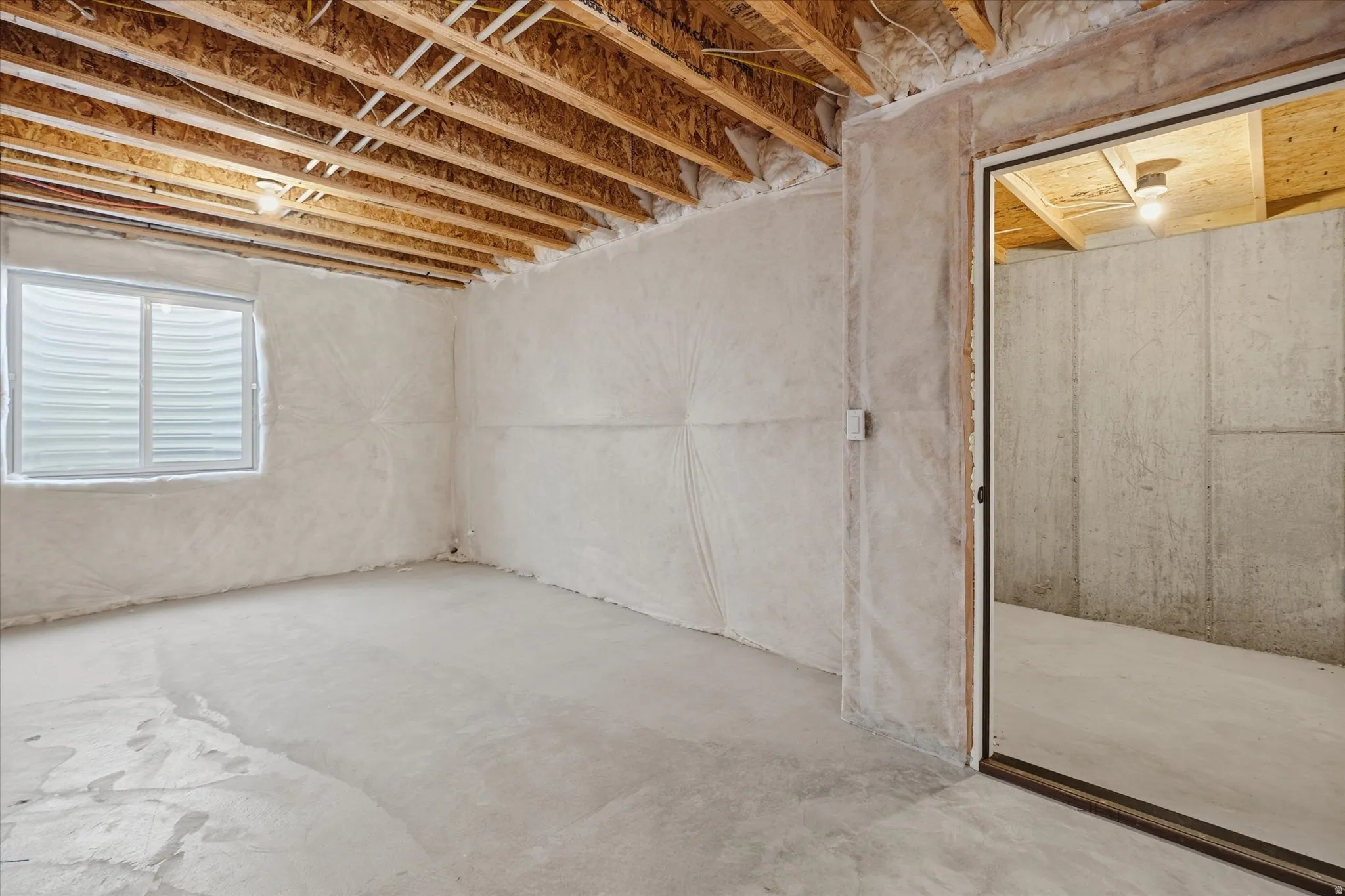 Unfinished basement featuring exposed wood joists, a single window with blinds, concrete flooring, and exposed concrete walls