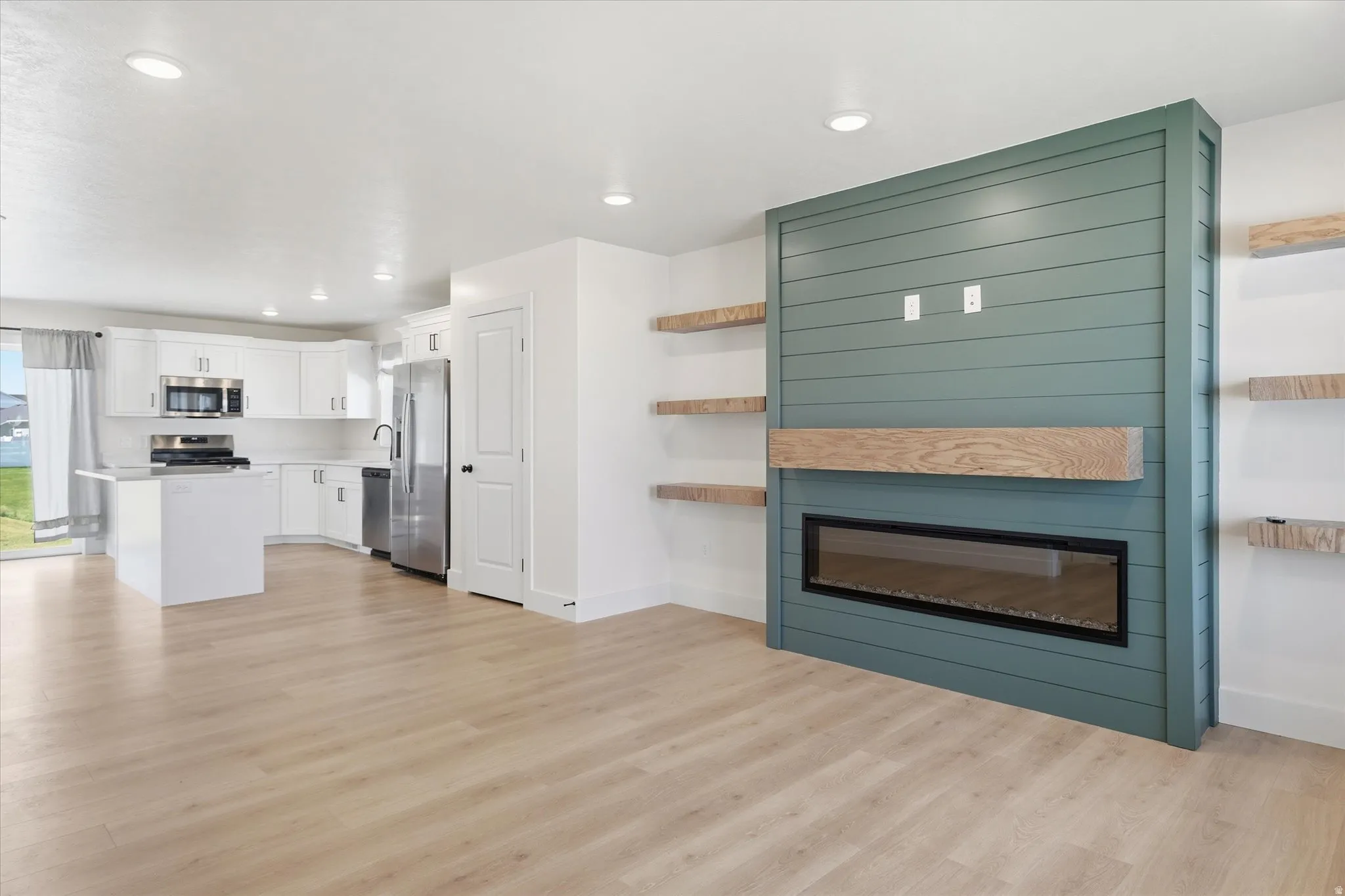 Light wood-finish flooring throughout, modern electric fireplace with a wood mantel, built-in shelving, white cabinetry with stainless steel appliances, and an island with a cooktop