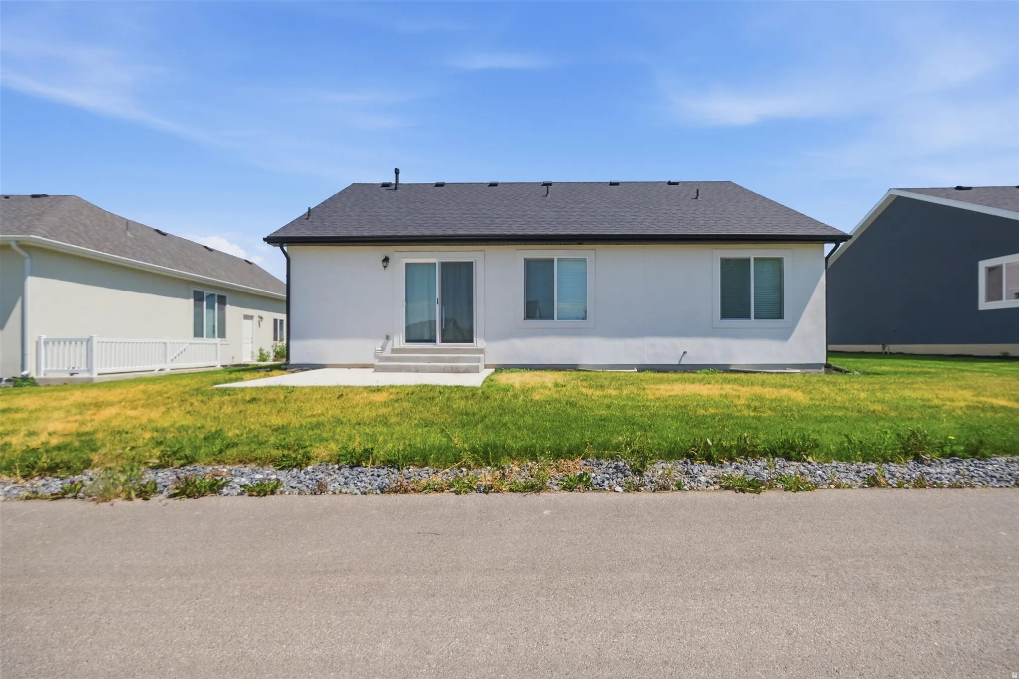 White stucco exterior with a dark shingle roof, featuring a sliding glass door, two windows, and a concrete patio