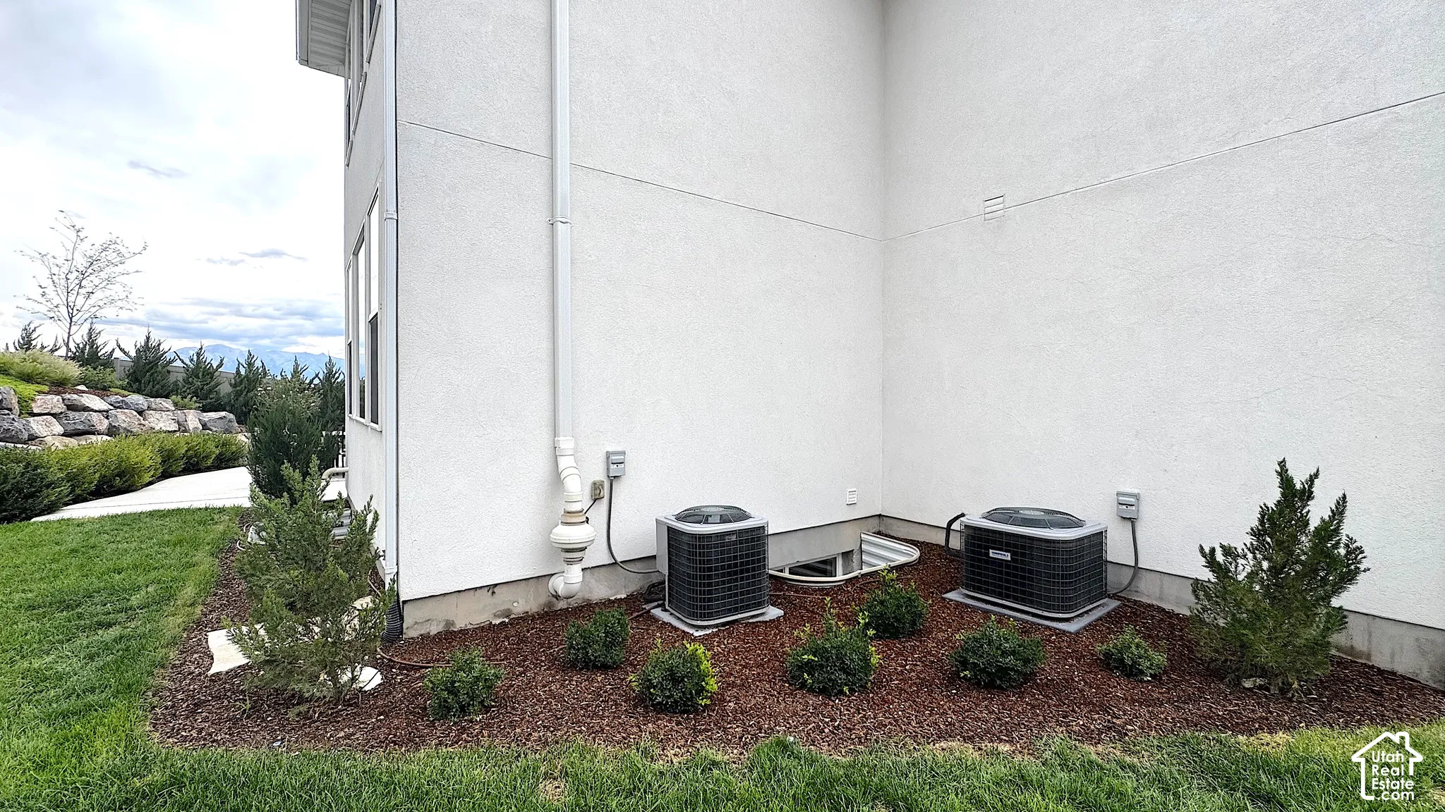 Stucco exterior with a landscaped perimeter, featuring two air conditioning units, decorative shrubs, and a sloped rock retaining wall