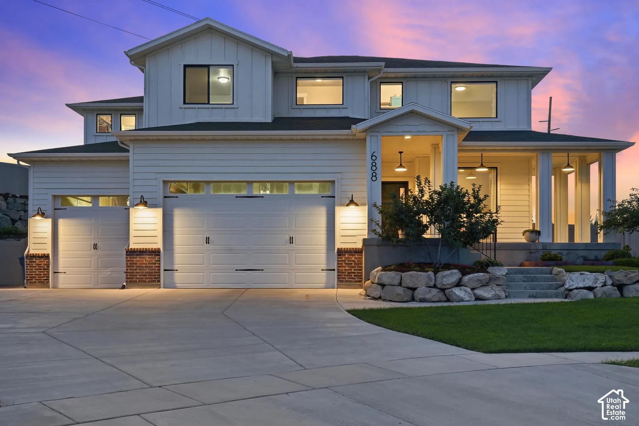 Modern farmhouse exterior featuring a covered porch, multiple garages, and a stone-edged landscape