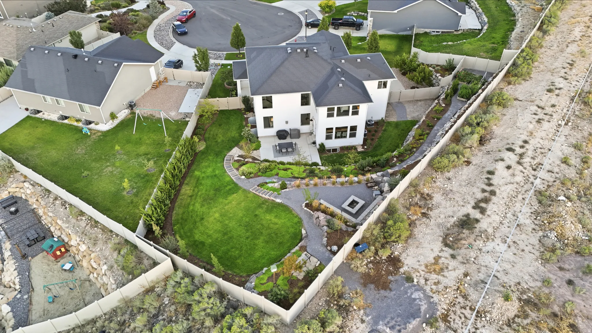 White exterior with dark roof, expansive green lawn, built-in fire pit, paved patio, and mature landscaping