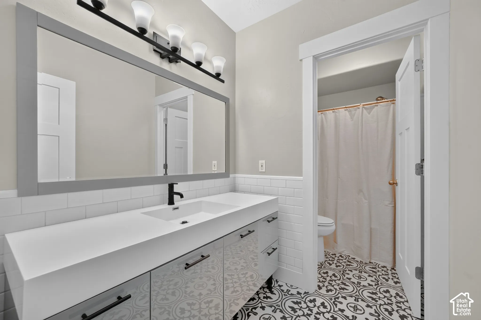 Bathroom featuring a single vanity with integrated sink, matte black faucet, wall-mounted light fixture, full-width framed mirror, and decorative patterned floor tiles