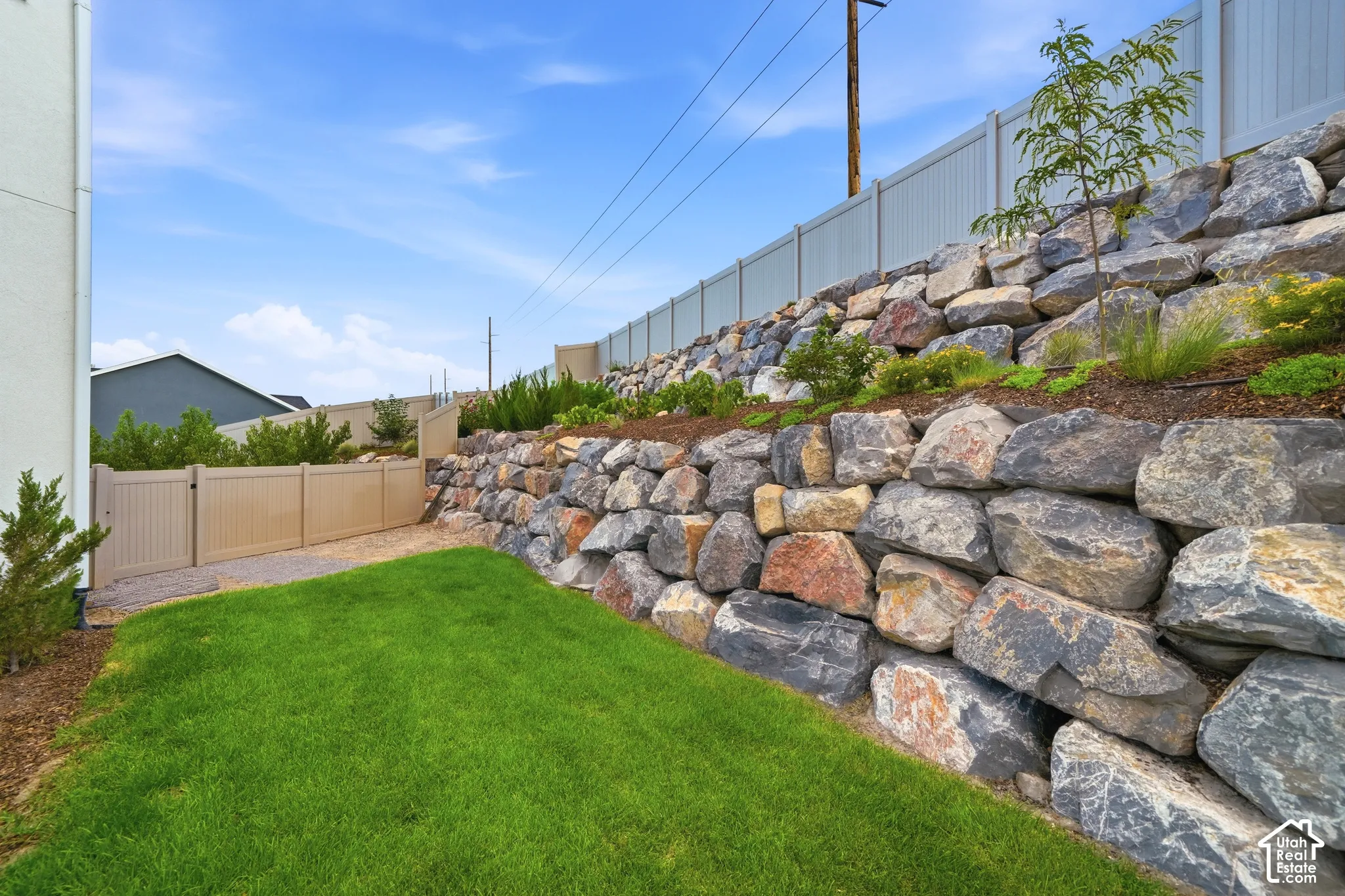 Manicured lawn with a large rock retaining wall, mature landscaping, and a privacy fence