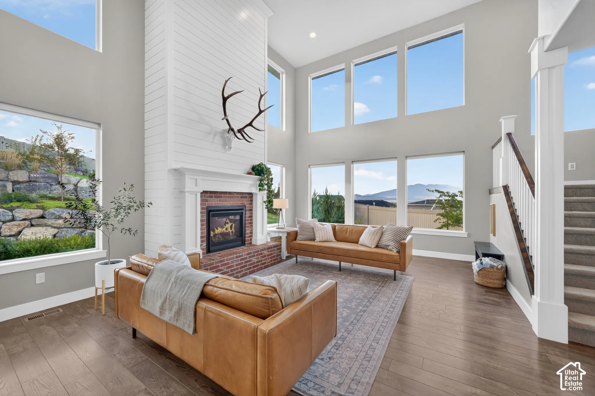 Two-story living area with high ceilings, a brick-surround fireplace, a shiplap accent wall, wood-finish flooring, and multiple large windows