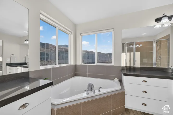 Built-in corner soaking tub with tile surround, dual windows, separate vanities with dark countertops, and a clear glass shower enclosure