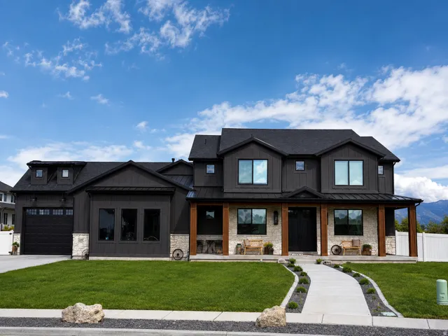 Modern farmhouse exterior with dark vertical siding, contrasting light stone accents, a covered front porch with substantial wood columns, a paved walkway, and an attached garage