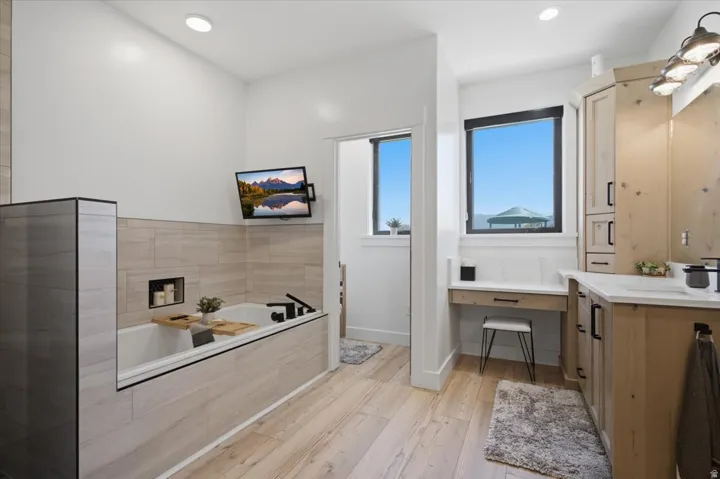Recessed soaking tub with integrated niche and surrounding tile, wood-finish flooring, built-in vanity with countertop, and dual windows