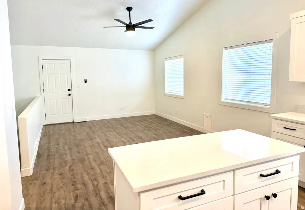 Vaulted ceiling with a ceiling fan, wood-finish flooring, multiple windows with blinds, and a kitchen island with white cabinetry and dark hardware